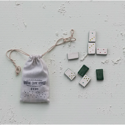 A set of ceramic dominoes with white, green, and dark gray tiles, displayed next to a small white drawstring pouch printed with 'HANDMADE CERAMIC DOMINOES' on a textured light gray background.