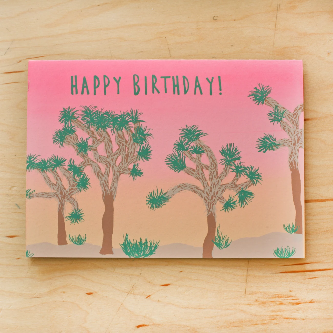 A blank greeting card with a hand-designed and screen printed pink and green joshua tree landscape on 100% cotton rag archival gray paper, including an envelope.