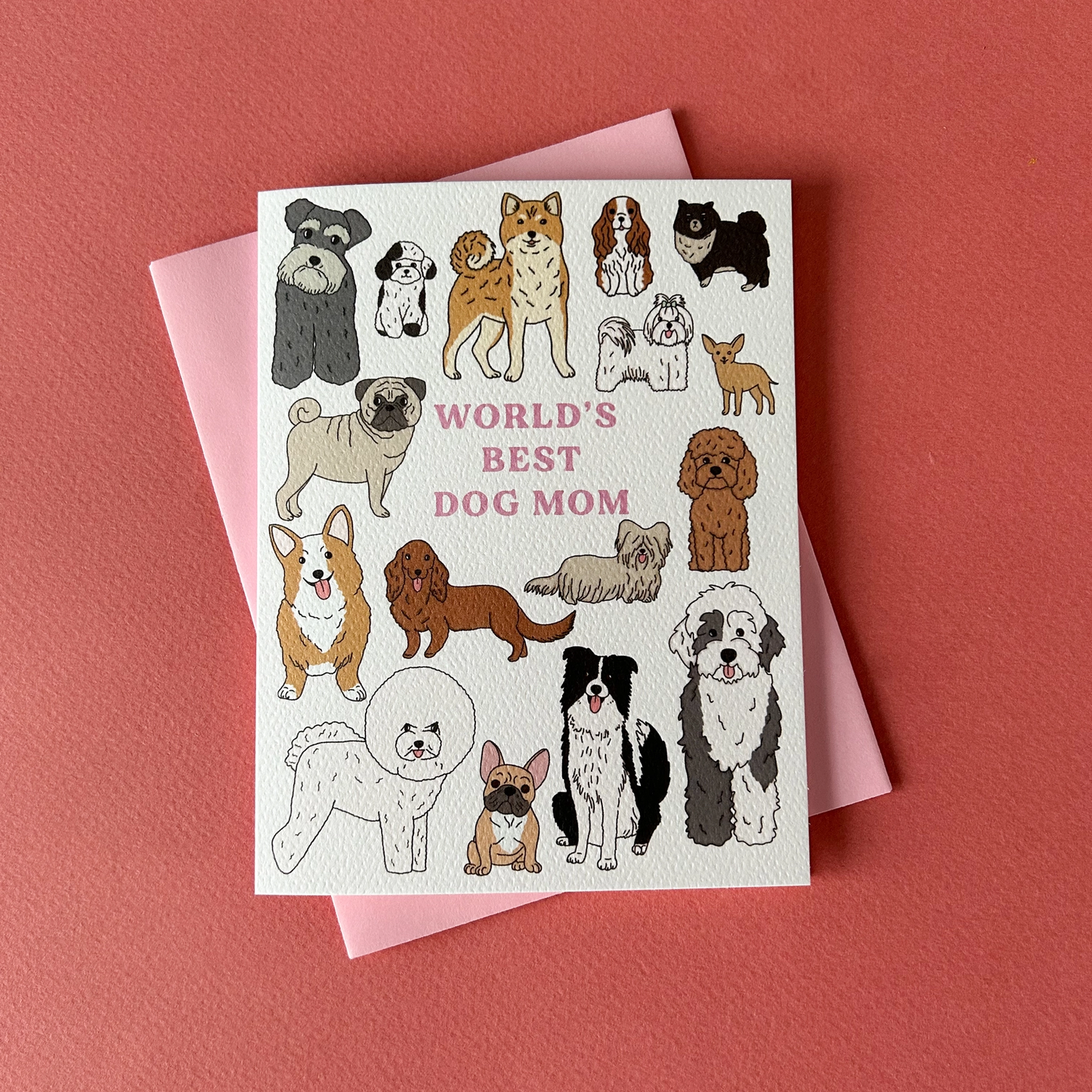 Greeting card with various illustrated dogs and the text 'WORLD'S BEST DOG MOM' across the top, accompanied by a pink envelope, all placed on a red background.