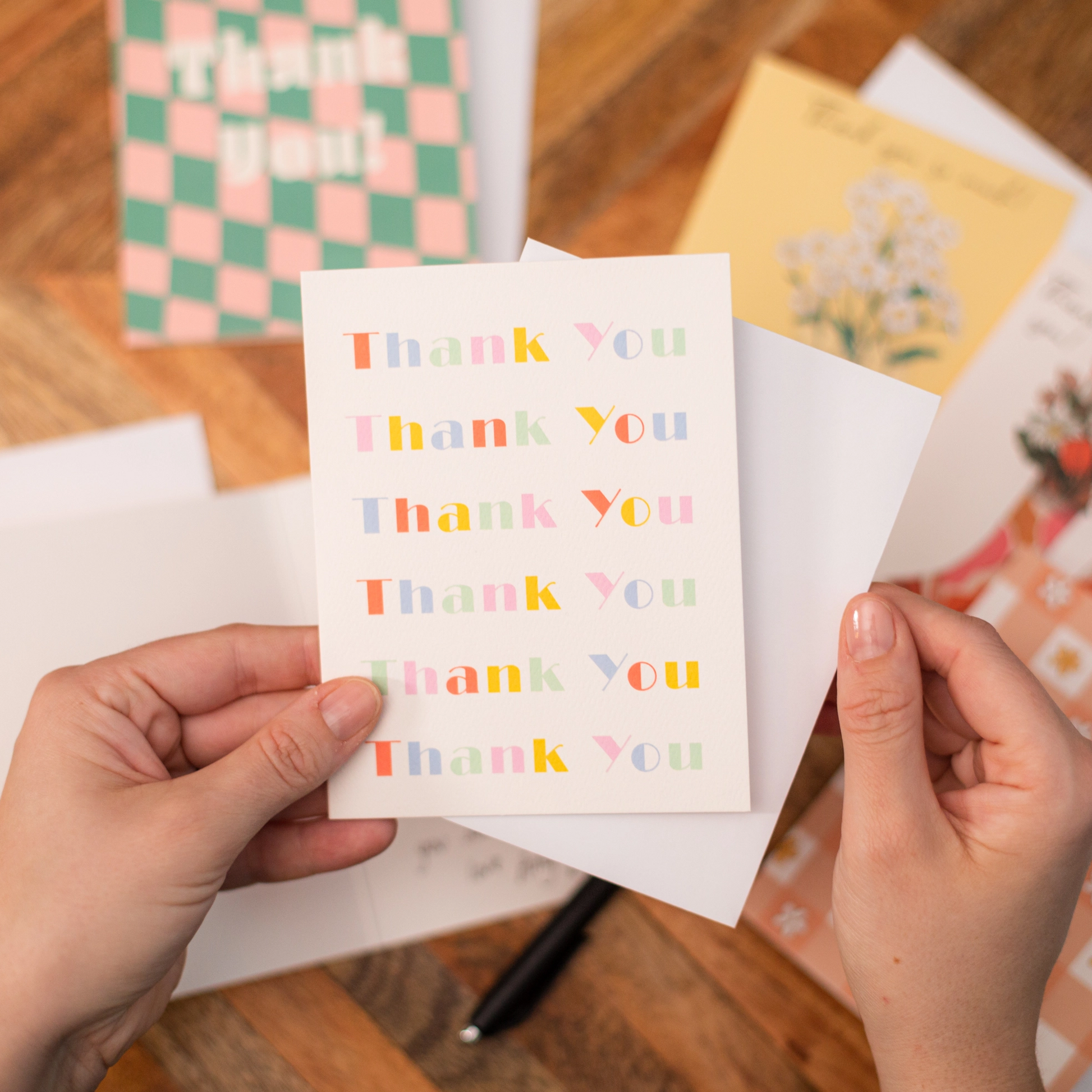 Person holding a 'Thank You' card with colorful text on a wooden surface with other cards in the background.