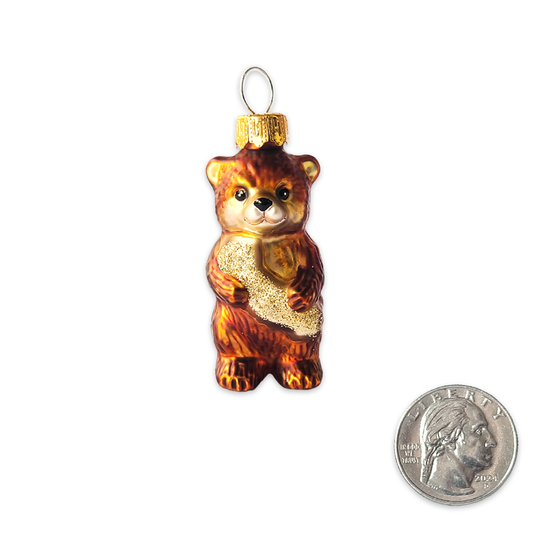 A small brown teddy bear glass ornament holding a gold glittery heart, shown next to a US quarter for scale on a white background.