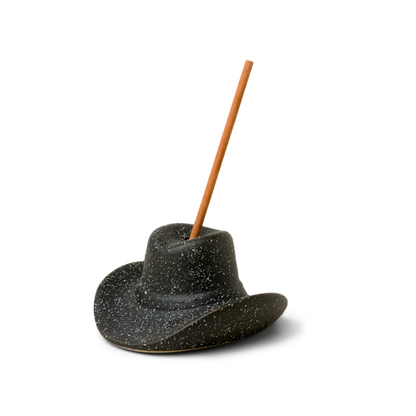 A black ceramic incense holder shaped like a cowboy hat with a stick of incense inserted on top.